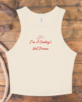 An off-white sleeveless tank top with red cursive text reading 'I'm A Cowboy's Wet Dream' and a small heart detail, laid flat on a rustic wooden background with rope and wood props.