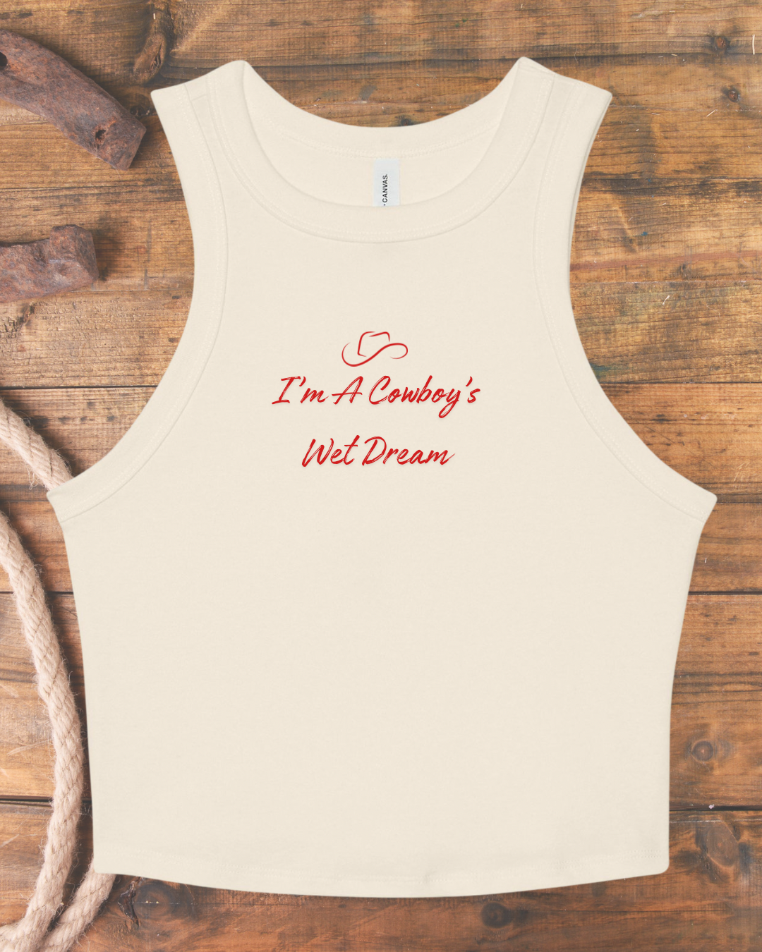 An off-white sleeveless tank top with red cursive text reading 'I'm A Cowboy's Wet Dream' and a small heart detail, laid flat on a rustic wooden background with rope and wood props.
