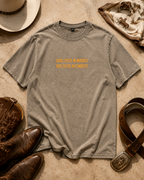 A washed brown gray t-shirt with orange text reading 'GOOD TASTE IN WHISKEY AND TASTE IN COWBOYS' laid flat on a textured beige surface, surrounded by cowboy boots, a hat, a bandana, and a leather belt.