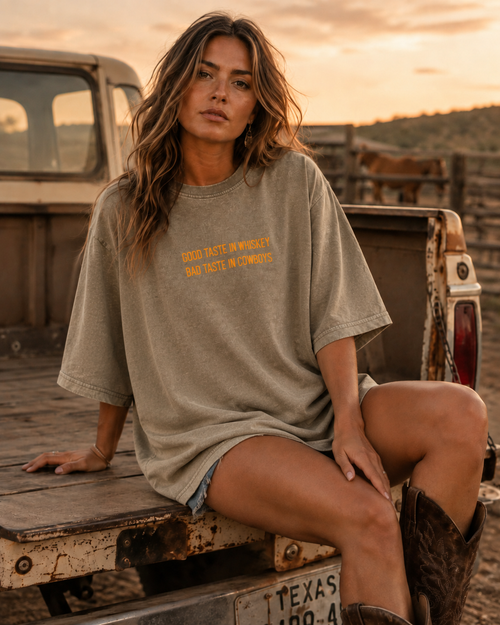 A woman sits in the bed of a rustic pickup truck at sunset, wearing an oversized faded taupe graphic t-shirt with orange text that reads 'GOOD TASTE INVENTED BY VASTE IN COWBOYS'.
