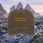 A brown baseball cap with yellow embroidered text reading 'GOOD TASTE IN WHISKEY BAD TASTE IN COWBOYS' is centered against a scenic mountain landscape background.