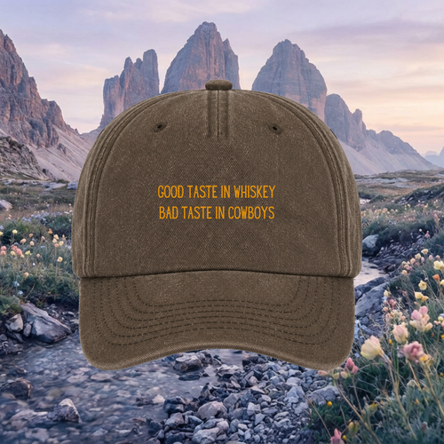 A brown baseball cap with yellow embroidered text reading 'GOOD TASTE IN WHISKEY BAD TASTE IN COWBOYS' is centered against a scenic mountain landscape background.