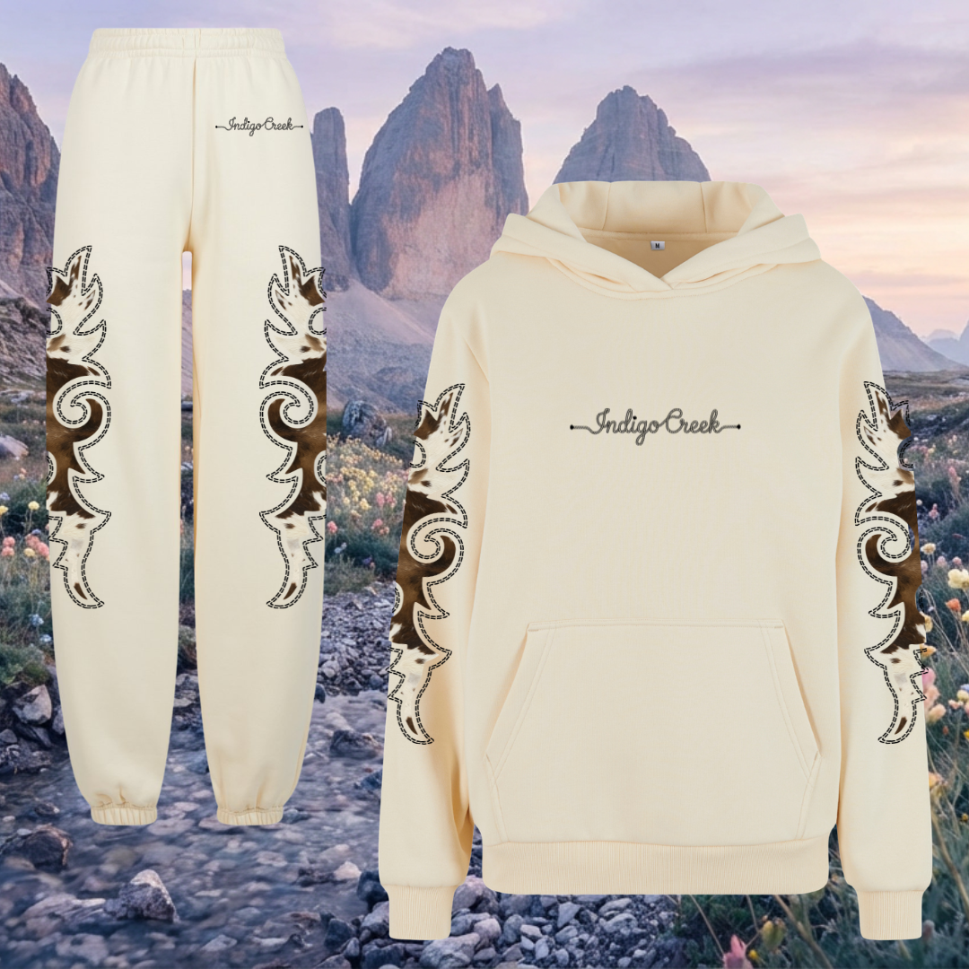 Cream-colored hoodie and sweatpants with decorative boot stitch and cow print patterns and 'Indigo Creek' branding against a western mountainous landscape.