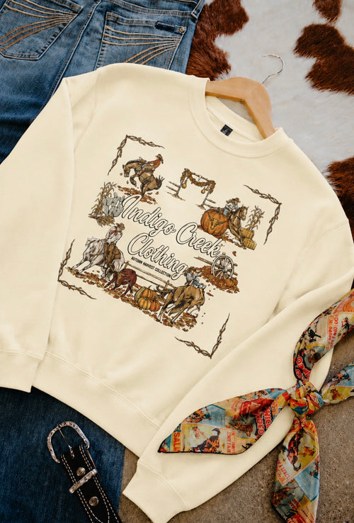 Cream-colored crewneck sweatshirt with a rustic western-themed graphic print, styled flat-lay with blue jeans, a patterned scarf, and a brown cowhide rug.