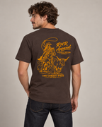 A man wearing a brown short-sleeve t-shirt with a large yellow graphic print on the back, featuring a cowboy and steer design with the text 'RNR Apparel' and 'The Cowboy West'.