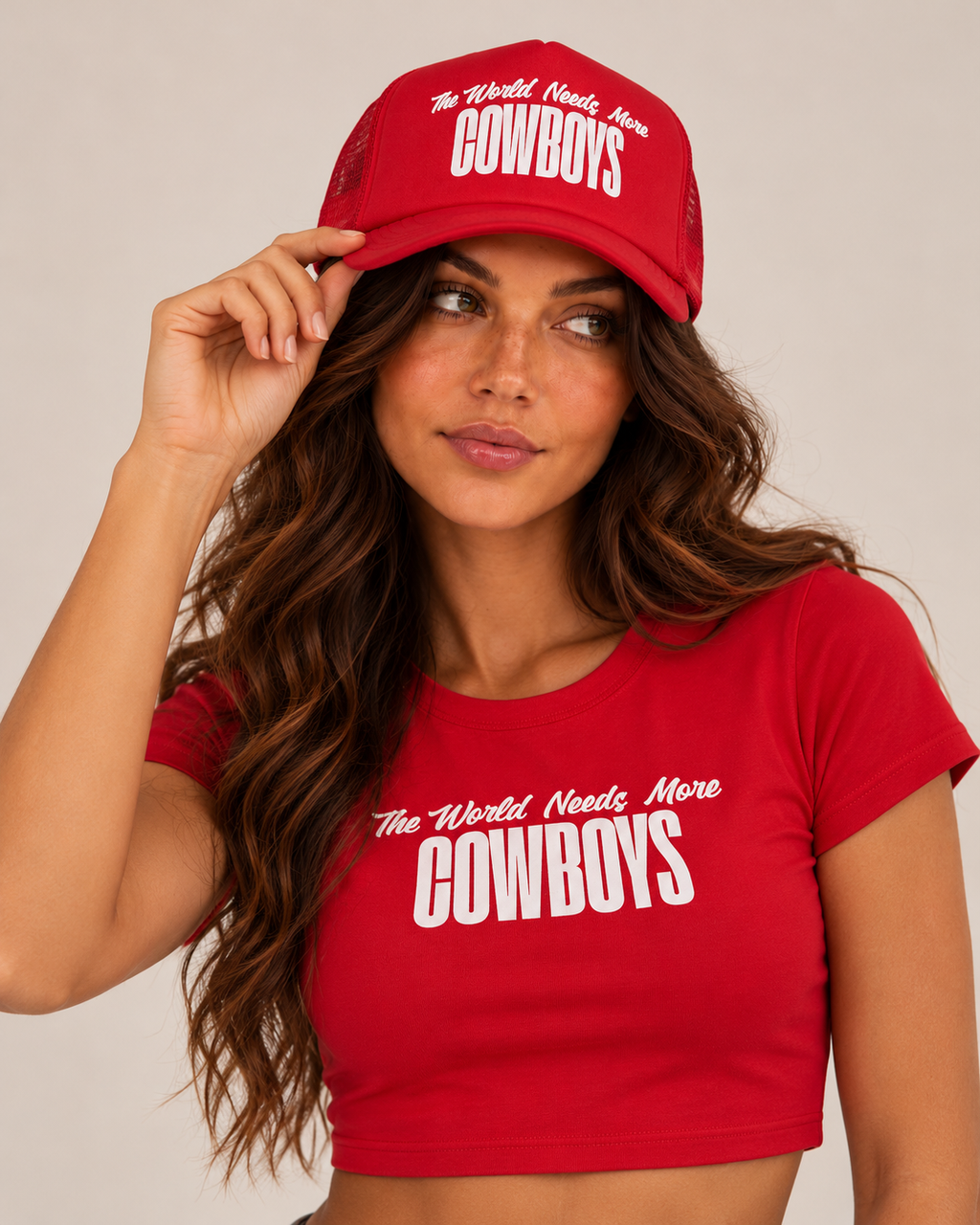 A woman wearing a red crop top and matching trucker hat, both printed with the phrase 'The World Needs More COWBOYS' in white text, against a plain light beige background.
