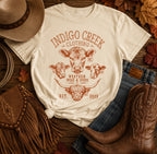 An off-white graphic t-shirt featuring a brown Western-themed design with three cow illustrations and the text 'INDIGO CREEK CLOTHING' and 'WESTERN WEAR & GOODS'. The shirt is styled in a flat-lay arrangement with a brown hat, blue jeans, brown leather cowboy boots, a brown fringe bag, and scattered autumn leaves on a rustic wooden background.