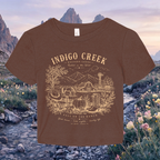 A brown short-sleeve t-shirt with a tan graphic print reading 'INDIGO CREEK' and 'RICHES OF THE VALLEY' above a farm illustration, shown against a scenic mountain and stream background.