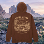 A brown pullover hoodie shown from the back, featuring a large tan graphic print with the text 'INDIGO CREEK' and an illustrated mountain-and-rustic-scene design. The hoodie is centered against a scenic outdoor background with rocky mountains and wildflowers.