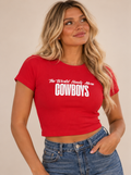 A woman wearing a bright red short-sleeve crop top with white text that reads 'The World Needs More COWBOYS', paired with blue jeans, against a plain beige background.