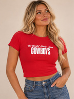 A woman wearing a bright red short-sleeve crop top with white text that reads 'The World Needs More COWBOYS', paired with blue jeans, against a plain beige background.