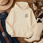 Cream-colored pullover hoodie with a small embroidered logo on the left chest, styled in a flat lay with jeans, a cowboy hat, boots, and a bandana on a wooden background.