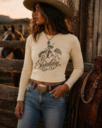 A woman wearing a cream long-sleeve crop top with a black western graphic that reads 'Sunday Radio Club,' paired with blue jeans and a cowboy hat, standing outdoors beside a rustic wooden fence and a horse.