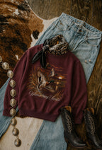 A styled flat-lay featuring a maroon sweatshirt with a duck hunting graphic and the text 'Wandering Souls'. The sweatshirt is styled with light-wash blue jeans, black cowboy boots, a black and brown bandana, and a silver coin necklace on a rustic wood and cowhide background.