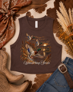 A sleeveless brown tank top laid flat on a cowhide surface, featuring a printed duck hunting scene with three mallards at sunset and the text 'Wandering Souls.' The top is styled with cowboy boots, blue jeans, a brown belt, a rust scarf, and dried pampas grass.