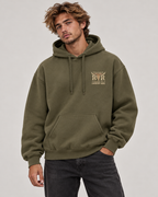 A man wearing an olive green pullover hoodie with a beige graphic on the left chest that reads 'Rustler Ranch' and 'COWBOY WEE', paired with dark gray jeans, against a light gray studio background.